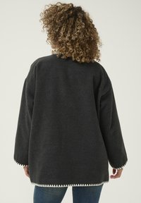 Överstor mörkgrå jacka gjord av mjukt tyg, med vit zigzag-söm längs fållen och ärmarna. Löst passande, långa ärmar.