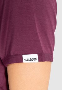 Chemise à manches courtes de couleur bourgogne profond, en tissu doux. Présente une étiquette blanche avec "SMILODOX" sur l'ourlet de la manche.