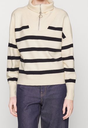 Kremfarvet sweater med sorte horisontale striber og halvlange lynlås krave, båret med mørkeblå jeans, vist fra skuldre til hofter.
