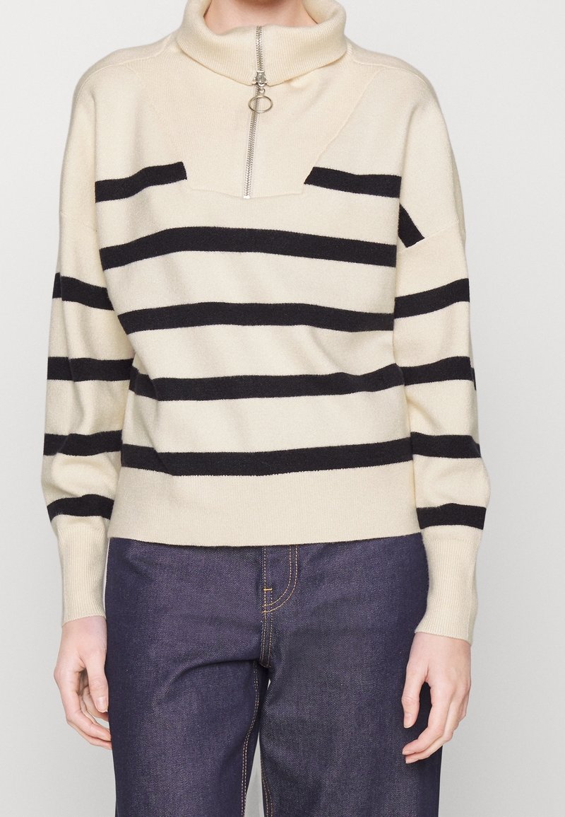 Pull crème avec des rayures horizontales noires et un col à demi-fermeture éclair, porté avec un jean bleu foncé, visible des épaules jusqu'aux hanches.