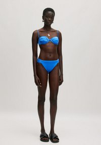 Blauer Bikini mit gerafften Details, Unterbrustbügel-Oberteil und verstellbaren Trägern; kombiniert mit passender, niedrig sitzender Unterhose. Das Modell trägt schwarze Slides.