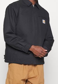 Černá bunda na zip s límcem, vybavená kapsou na hrudi a nášivkou s logem. Vyrobena z hladké, odolné látky s zesílenými švy.