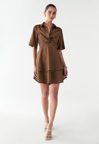 Rjava obleka s kratkimi rokavi z vozlom na prsih, plastenjski krilo in gladko teksturo. Združena z belimi sandali na vezalke.