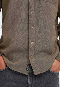 Bruine textuurshirt met een knoopsluiting aan de voorkant, met lichtbruine knopen en een borstzak. Gepaard met donkere jeans en een label aan de zoom.
