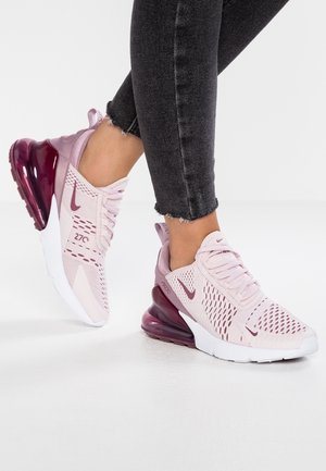 Person iført lyserøde og burgundy Nike Air Max 270 sneakers og sorte korte jeans står på en hvid gulv.