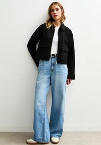 Veste noire cropped avec deux poches avant, portée sur un haut blanc et un jean large bleu clair, associée à des baskets beiges.