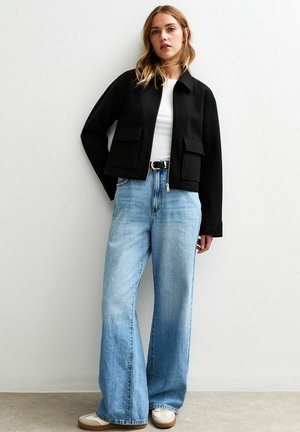 Svart kort jacka med två framfickor, bärs över en vit topp och ljusblå vida jeans, i kombination med beige sneakers.