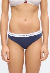 Femme portant un slip bikini bleu marine avec une ceinture élastique blanche marquée "Tommy Hilfiger" et un soutien-gorge de sport blanc avec un petit logo.
