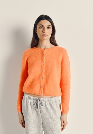 Felle oranje fluffy cardigan met een ronde halslijn en een sluiting met zeven knopen aan de voorkant. Draagt over grijze sweatpants met een elastische tailleband.