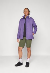 Purple waterproof jacket with a zip front and hood, worn over a black top, paired with olive green shorts and black sneakers.