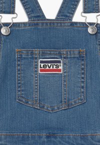 Džínové lacláče ve středně modré barvě s kovovými komponenty stříbrné barvy. Obsahují nastavitelné ramínka, přední kapsu s logo nášivkou Levis a kontrastní švy.