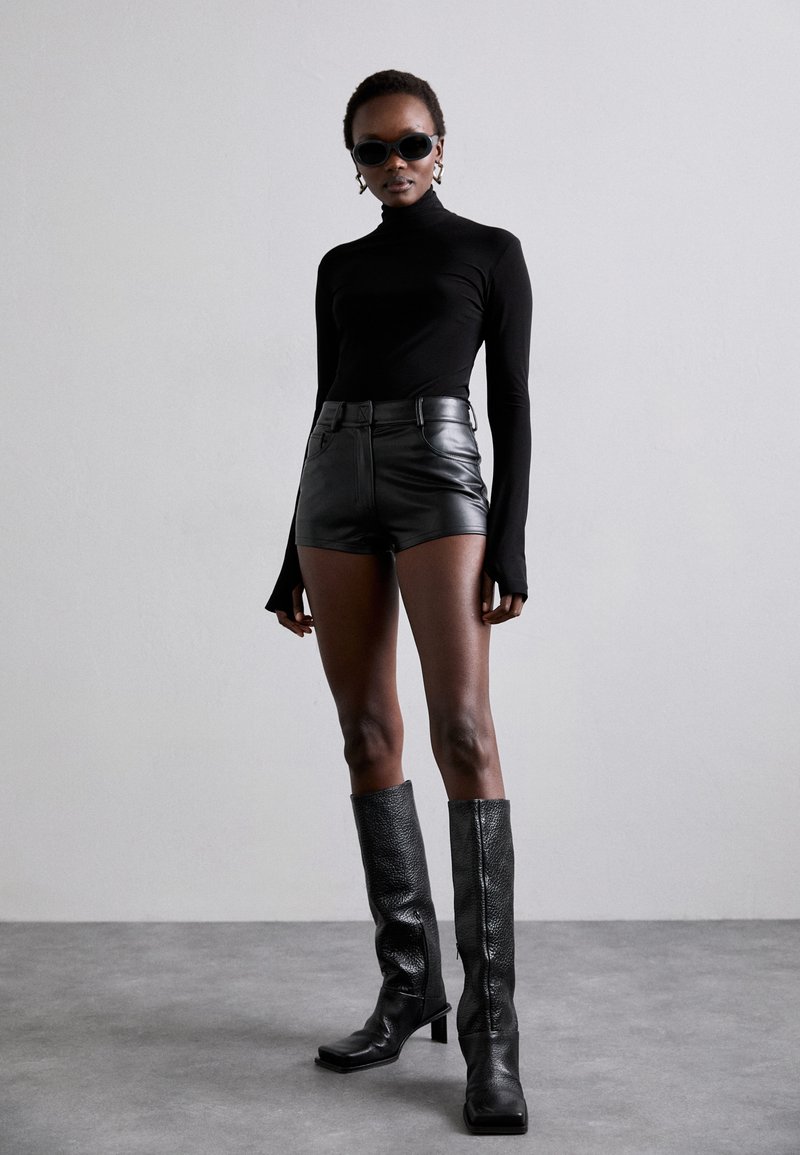 Camiseta de cuello alto negra, pantalones cortos de cuero negros de cintura alta, botas negras con textura hasta la rodilla y gafas de sol negras. Diseño simple y elegante.
