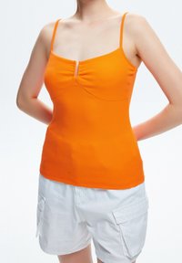 Femme portant un haut orange ajusté sans manches avec de fines bretelles et un short cargo blanc, les bras détendus derrière le dos, sur un fond uni.