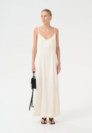 Langes, cremefarbenes Satin-Kleid mit dünnen Trägern, V-Ausschnitt, seitlichen Schlitzen und einer lässigen Silhouette, kombiniert mit einer kleinen schwarzen Handtasche und Sandalen.