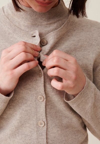 Cardigan en tricot beige clair avec un col droit haut et cinq boutons, tenu par des mains en boutonnant le haut. Texture douce, détails minimes.
