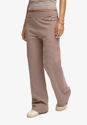 Frau trägt braune und beige gestreifte, weite Hosen, beige Sneaker und ein passendes gestreiftes Oberteil, steht vor weißem Hintergrund.