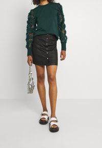 Woman wearing a green top with sheer floral sleeves, black button-front leather mini skirt, white sandals, holding a small zebra-print handbag.