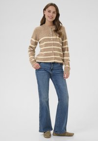 Gaiši brūns adīts kardigāns ar krēmkrāsas svītrām, apaļš dekoltē un priekšējie pogas, kombinēts ar ziliem platiem džinsiem un bēšiem mokasīniem.