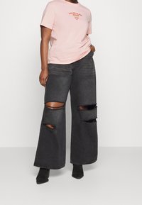 Černé široké džíny s roztrhanými koleny, spárované s lehkým růžovým bavlněným tričkem s oranžovým logem. Roztrhaná textura na džínách.