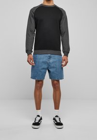 Personne portant un sweat-shirt à manches longues noir et gris foncé, un short en jean bleu, des chaussettes blanches et des baskets noir et blanc, debout sur un sol gris.