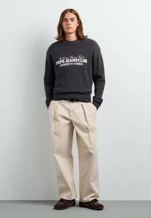 Giovane uomo con i capelli lunghi che indossa un maglione Pepe Jeans Club grigio scuro, pantaloni beige a gamba larga e scarpe marrone scuro, in piedi con le mani nelle tasche.