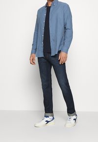 Jasnoniebieska koszula z guzikami, ciemne dżinsy i białe sneakersy z niebieskimi akcentami. Gładki materiał, luźny krój i podwinięte mankiety spodni.