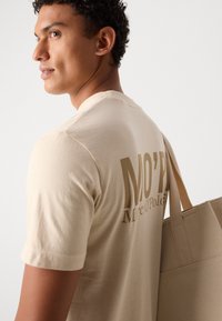 Beige cotton t-shirt with short sleeves, featuring large dark beige text "MO'P" and "Marc O'Pol." Model holds a textured tan tote bag.