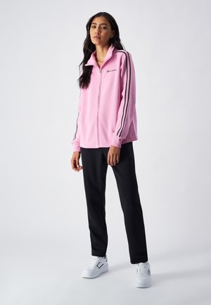 Felpa rosa con zip, strisce nere laterali e logo; abbinata a pantaloni neri e sneakers bianche. Tessuto morbido, vestibilità rilassata.