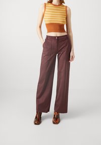 Pletený crop top v oranžové a žluté barvě s texturovaným vzorem; široké vínové kalhoty s bočními kapsami; lesklé hnědé boty s hranatými špičkami.