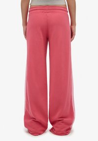 Persona che indossa pantaloni della tuta rosa larghi con elastico in vita, in piedi con le mani ai fianchi, visibile dalla vita in giù, su sfondo bianco.