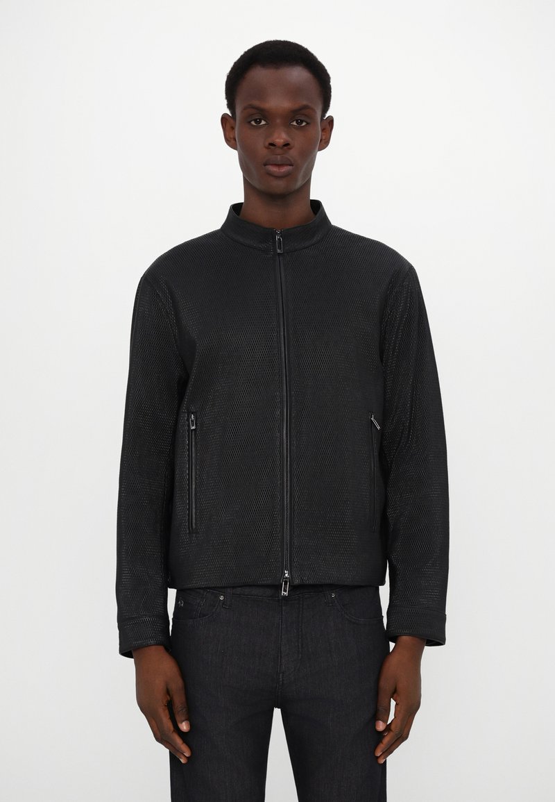 Schwarze Zip-Jacke aus strukturiertem Netzgewebe. Verfügt über zwei mit Reißverschluss gesicherte Fronttaschen und einen Stehkragen. Trägt man über dunklen Jeans.