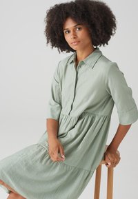 Hellgrünes, strukturiertes Hemdkleid mit geknöpftem Vorderteil, kragenlosem Ausschnitt und Dreiviertelärmeln. Ausgestellter, gestufter Rock; lässiges Design.