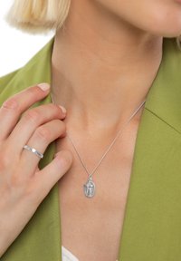 Sidabrinė pakabuko vėrinys su ovaliu pakabuku, kuriame yra sudėtingų detalių, dėvimas ant plonos grandinėlės priešais žalią švarką.