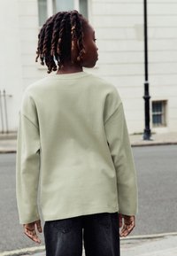 Šviesiai žalia, tekstūruota ilgomis rankovėmis palaidinė su nuleistais pečiais, derinama su tamsiais džinsais, matoma iš nugaros gatvės fone.