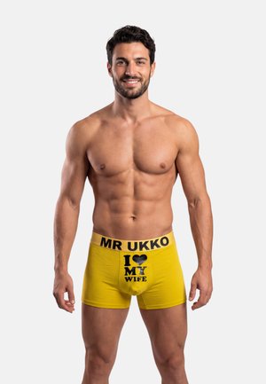 Hombre sin camiseta con cabello corto y oscuro y barba, vistiendo calzoncillos bóxer amarillos con la cintura que dice "MR UKKO" y texto en el frente que dice "I [corazón] MI ESPOSA".