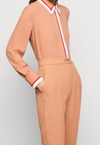 Completo in pesco con maniche lunghe, colletto a righe bianche e rosse, vita con cinturino, tessuto liscio e un accento dorato in vita.