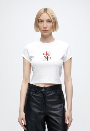 Joven mujer con el pelo rubio corto que lleva una camiseta blanca corta de Calvin Klein y pantalones de cuero negros sobre un fondo blanco liso.
