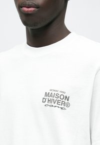 Lähivaade inimesest, kes kannab valget dressipluusi, mille rinnal on musta tekstiga "MCMXC 1990 MAISON D'HIVER core".