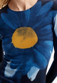 Top de manga larga acanalado en azul oscuro con un gran estampado de girasol que presenta un centro amarillo y pétalos en azul claro. Textura suave.