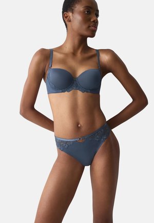 Femme debout, mains derrière le dos, portant un soutien-gorge rembourré bleu et une culotte assortie bordée de dentelle sur fond uni.