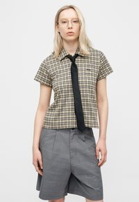 Νεαρό άτομο με μακριά ξανθά μαλλιά, φορά γυαλιά, μια μπεζ καρό κοντομάνικη μπλούζα με μαύρη γραβάτα και γκρι καρό σορτς μέχρι το γόνατο.