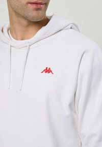 Mees valges kapuutsiga, millel vasakul rinnal on punane Kappa logo tikitud, näitab alahuult ja kaela neutraalse tausta ees.