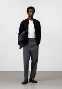 Svart bomberjacka, vit skjorta och grå byxor. Slät läderväska med texturerad yta. Svarta slipskor med minimalistisk design.