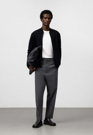 Giubbotto bomber nero, camicia bianca e pantaloni grigi. Borsa in pelle liscia con finitura testurizzata. Scarpe slip-on nere dal design minimale.