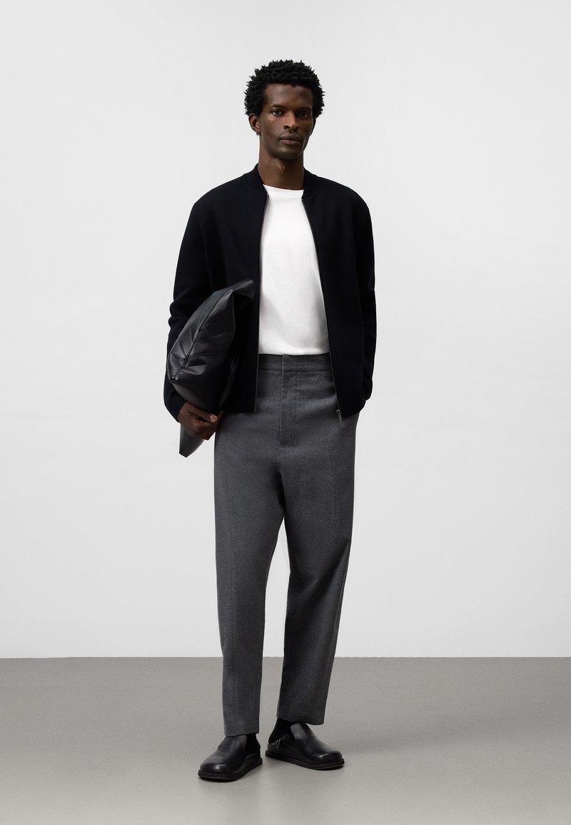 Svart bomberjacka, vit skjorta och grå byxor. Slät läderväska med texturerad yta. Svarta slipskor med minimalistisk design.