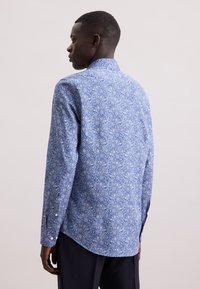Uomo di colore di profilo a sinistra, che indossa una camicia a maniche lunghe blu con motivo floreale e pantaloni scuri, su uno sfondo semplice.