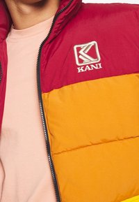 Prošívaná bunda s horní částí v barvě tmavě červené a spodní částí v barvě žluté, se zipem a vyšitým logem "KANI" na hrudi.