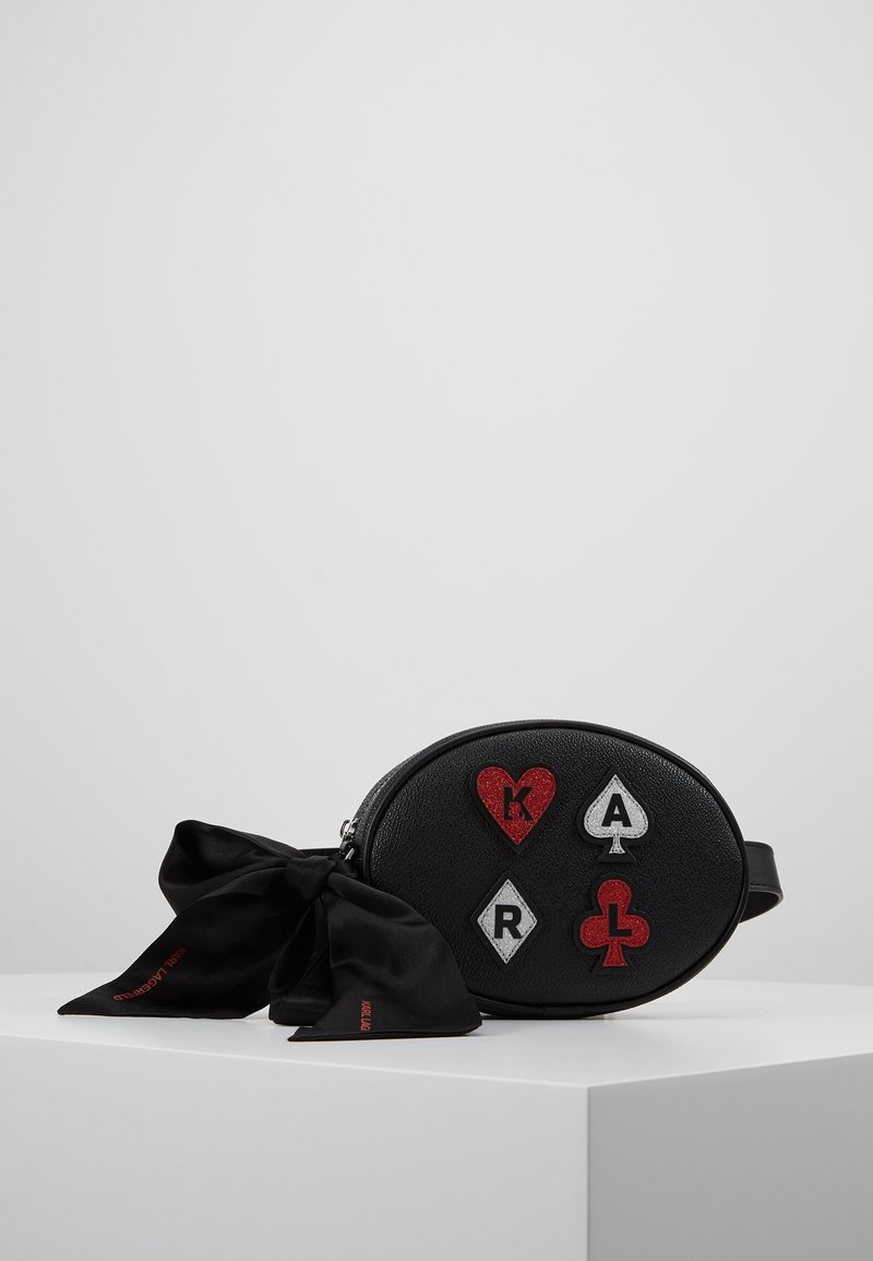 Sac en cuir ovale noir avec des patchs rouges et blancs aux motifs de couleurs de cartes à jouer, et un nœud en tissu noir attaché sur le côté, reposant sur une surface blanche.