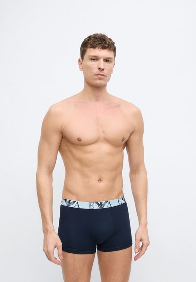 Άνδρας χωρίς πουκάμισο στέκεται, φορώντας σιδερένιες κοντές μπλούζες ναυτικού μπλε με ανοιχτό γκρι ζώνη μέσης που φέρει επαναλαμβανόμενο μαύρο λογότυπο και γράμματα.