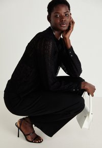 Haut à manches longues en dentelle noire associé à un pantalon noir à pattes d'éléphant et des escarpins noirs à lanières. Tenant un petit sac à main blanc. Fond neutre.